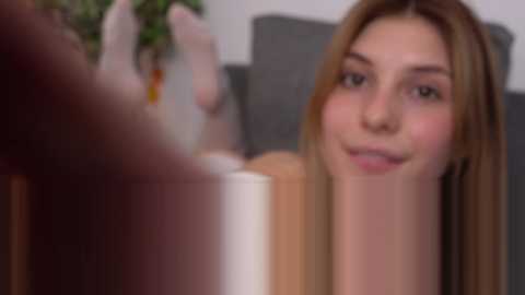 Media: A blurred video of a young woman with light skin and long blonde hair, wearing a white shirt, smiling, with a blurred background of a room and a potted plant.