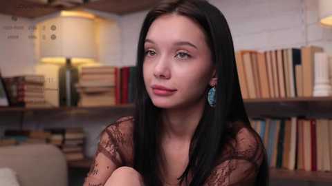 Media: Video of a young woman with fair skin and long black hair, wearing a sheer black lace top, sitting in a cozy, book-filled room with a warm lamp and beige walls.