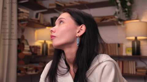 Media: Video of a pale, long-haired woman with dark eyes, wearing a beige sweater and blue earrings, looking contemplative in a cluttered, bookshelf-filled room with lamps and potted plants.