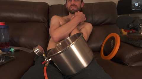 Media: Video of a shirtless man with a beard and a pot on his head, sitting on a brown leather couch. Background includes a laptop, orange Frisbee, and a blue water bottle.