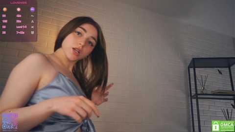 Media: Video of a young woman with long brown hair, wearing a blue dress, standing in a dimly lit room with a white brick wall and a black metal shelf in the background.