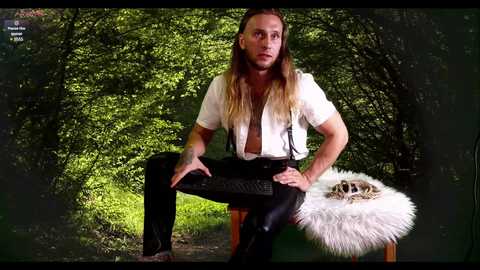 Media: Video of a muscular man with long blonde hair, wearing a white shirt, black pants, and suspenders, sitting on a chair in a lush, green forest.