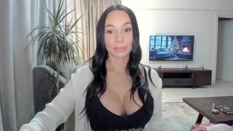 Media: Video of a light-skinned woman with long, black hair, wearing a black dress and white cardigan, standing in a modern living room with a flat-screen TV, potted plants, and a fireplace.