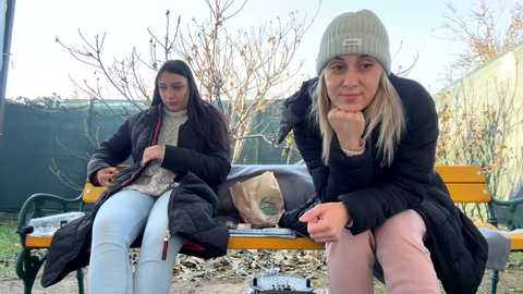 Media: Video of two young women, one with long dark hair and a beige beanie, the other with long blonde hair and a gray beanie, sitting on a park bench in winter attire, holding shopping bags.