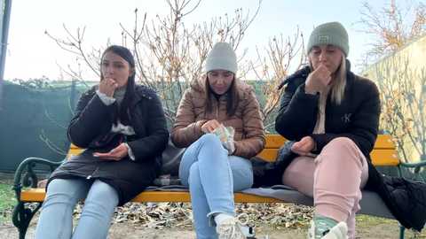 Media: Video of three young women sitting on a park bench, wearing winter clothes, hats, and holding cigarettes, with bare trees and a green fence in the background.