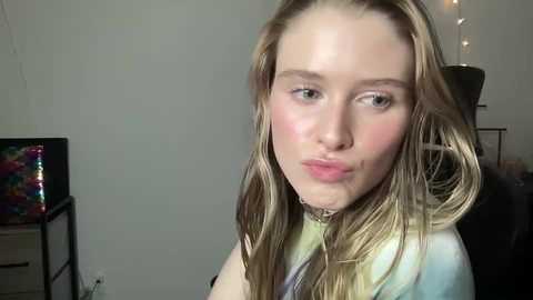 Media: Video of a young Caucasian woman with long, wet blonde hair, slightly blowing a kiss, wearing a tie-dye shirt. Background includes a colorful abstract painting, a lamp, and a dark-colored chair.