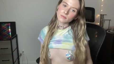 Media: Video of a young woman with long blonde hair, wearing a tie-dye crop top and a silver belly chain. She stands in a simple room with a colorful painting, a chair, and fairy lights in the background.