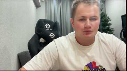 Media: Video of a man with short brown hair, wearing a white t-shirt, sitting in a gaming chair. Background features a Christmas tree and gray curtains.