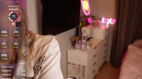 Media: A video of a dimly lit bedroom featuring a young woman with long blonde hair wearing a white hoodie, standing beside a mirror with a pink neon sign.