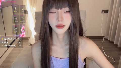 Media: Video of an Asian woman with long black hair, fair skin, and closed eyes, wearing a strapless lavender top. The room has beige walls, a gray couch, and a TV showing a digital clock.