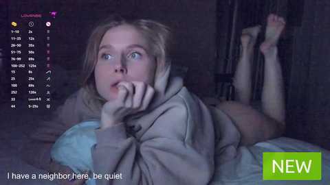 Media: Video of a blonde woman lying on her stomach in a dimly lit room, wearing a light-colored hoodie, legs raised, blue bedding, and text overlay reading \"I have a neighbor here be quiet.\