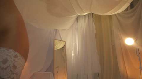 Media: Video of a romantic bedroom with sheer white curtains and soft lighting, creating a cozy, intimate ambiance. The bed is adorned with white bedding and a bouquet of white flowers on the left side.