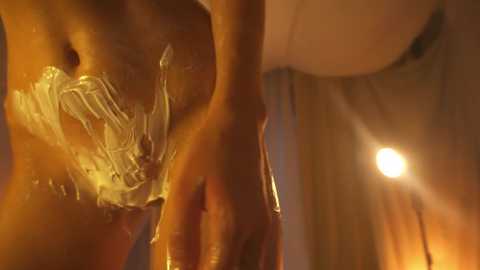 Media: Video: Close-up of a nude, light-skinned woman with a glistening body, covered in white, viscous liquid, leaning against a warm, lit wall. Her hand is on her hip.