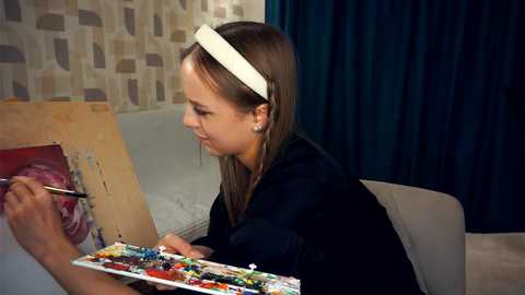 Media: Video of a young girl with straight brown hair, wearing a white headband, painting on a canvas. She's in a cozy room with beige patterned wallpaper and dark blue curtains. She holds a paintbrush in her right hand.