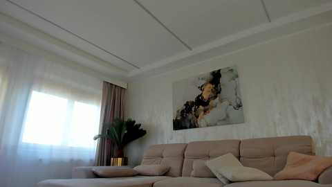 Media: Video of a minimalist living room with a beige sectional sofa, light-colored walls, large abstract painting, sheer white curtains, and a potted plant.