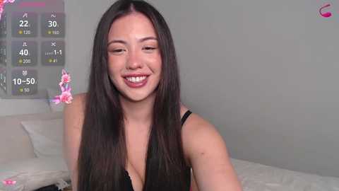 Media: Video of a smiling, fair-skinned, long-haired woman with medium-sized breasts in a black top, sitting on a bed with pink flowers in the background.