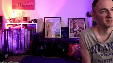 Media: Video of a man in a gray tank top, smiling, in a dimly lit room with purple and pink lighting. Behind him, vintage magazines and a dark couch.