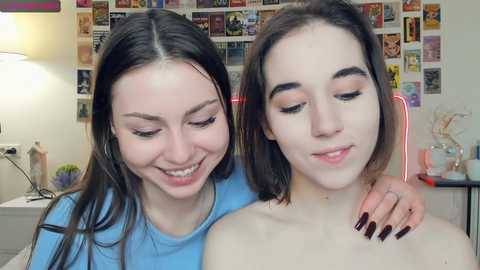 Media: Video of two young women with fair skin and dark hair, one with long hair and the other with shoulder-length hair, smiling and hugging in a cozy, colorful bedroom.