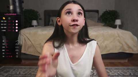 Media: Video of a young woman with long dark hair, wearing a white tank top, looking toward the camera with a neutral expression in a modern bedroom with a beige bedspread.