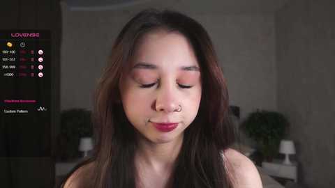 Media: A video of a young Asian woman with long dark hair, light skin, and closed eyes, wearing a nose ring. The background shows a dimly lit room with a plant and a lamp.