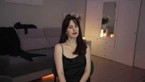 Media: Video of a woman with long dark hair and red lipstick, wearing a black camisole, seated in a modern living room with grey couch, white cabinets, and dim lighting.