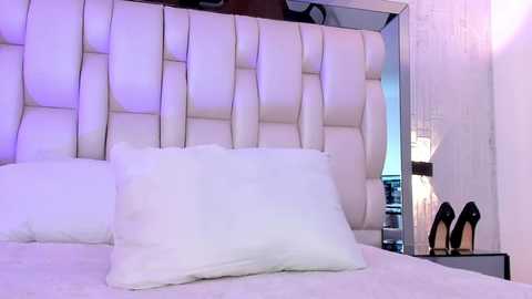 Media: Video of a modern bedroom with a white, tufted leather headboard, two white pillows, and a pair of black high heels resting on a bedside table. The room has a minimalist, luxurious aesthetic.