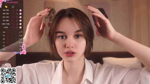 Media: A video of a young woman with fair skin and brown hair, wearing a white shirt, adjusting her hair in a bedroom with a bed and a blurred background.