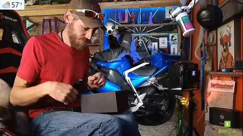 Media: Video of a bearded man in a red shirt and baseball cap, working on a blue motorbike in a cluttered garage, surrounded by tools and posters.