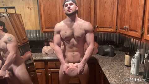 Media: Video of a muscular, naked man with a headband, sitting on a kitchen counter, holding a large, realistic-looking prosthetic penis. Background features wooden cabinets and countertop with kitchen items.