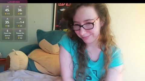 Media: Video of a young woman with glasses, long brown hair, wearing a turquoise t-shirt, smiling, lying on a bed with a Snorlax plush toy, in a room with a Pok\u00e9mon-themed wall calendar.