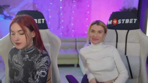 Media: Video of two women in white turtlenecks, one with red hair, one blonde, seated in gaming chairs with MASTERCARD logos, in a purple-lit, softly lit room.