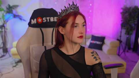 Media: Video of a fair-skinned, red-haired woman in a sheer black dress with a crown, seated in a beige gaming chair. Background includes a purple-lit room with plants and a couch.