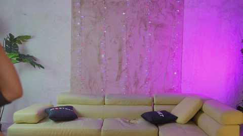 Media: Video of a modern living room with a beige sectional sofa adorned with black decorative pillows. A large, colorful, sparkly curtain hangs behind, illuminated by purple light.