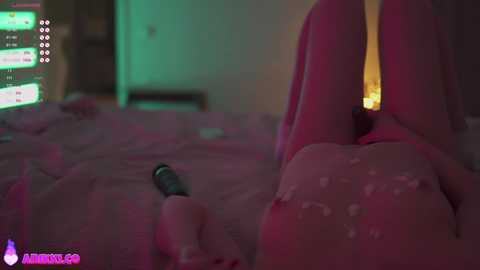 Media: Video of a nude woman with pink hair, lying on a bed with pink sheets, holding a black remote control, surrounded by a dimly lit room with green and pink lighting, and a television displaying various icons and text in the background.