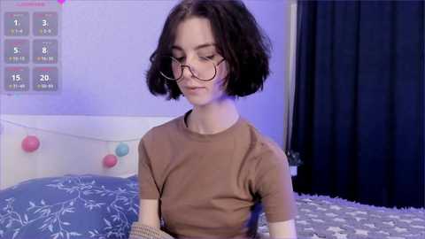 Media: A video of a young woman with short dark hair and glasses, wearing a brown top, sitting on a bed with white and blue patterned bedding, surrounded by pink and blue balloons.