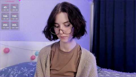 Media: Video of a young woman with short, wavy brown hair, wearing glasses, beige knit cardigan, and a brown shirt, in a bedroom with blue curtains and a bed with a white duvet.