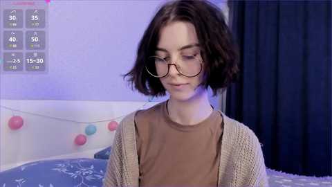 Media: Video of a young woman with short, dark hair, wearing glasses, a beige knit cardigan, and a brown top. Background features a bed with blue bedding, pastel balloons, and a digital display showing temperature and weather icons.