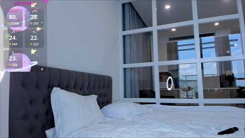 Media: Video of a modern bedroom with a gray tufted headboard, white bedding, and large windows. A smartphone screen with a virtual character overlaid on the left, indicating a virtual reality experience.