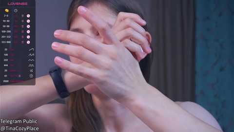 Media: A video of a woman with light skin and long brown hair covering her face, wearing a black smartwatch. The background features a dark curtain and a blue patterned wall.