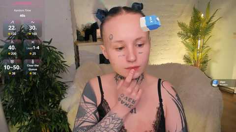 Media: A video of a fair-skinned woman with dark hair in two buns, wearing a black lace bralette and tattoos, sitting on a beige couch, with a plant and a blue box in the background.