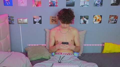 Media: Video of a shirtless young man with curly hair, wearing gray pants, sitting on a bed, texting on a phone, surrounded by colorful posters on a blue wall.