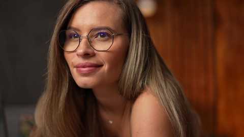 Media: Video of a smiling young woman with light skin, long straight blonde hair, and round glasses, set against a warm, blurred wooden background.