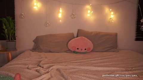 Media: A cozy bedroom scene with a plush pink pineapple pillow, beige bedding, fairy lights, and a green potted plant.