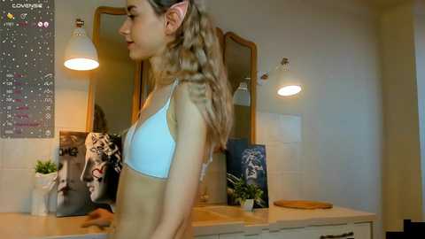 Media: A video of a young woman with long blonde hair in a light blue bra, standing in a modern bathroom with a calendar, plants, and mirrors.