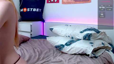 Media: Video of a young woman with light skin and long blonde hair lying on a bed with white pillows and a green leaf-patterned duvet. A black \"Mostbet\" sports jersey hangs on a wall behind her.