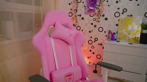 Media: Video of a pink, ergonomic gaming chair with white armrests and a logo, in a colorful, modern room with a white, patterned wall, a desk, and yellow artwork.