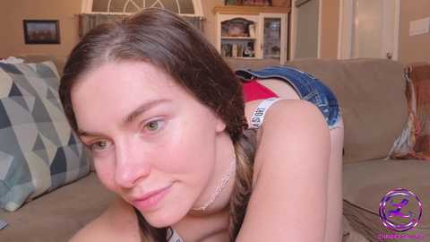 Media: Video of a young Caucasian woman with long brown hair, light skin, and green eyes, wearing a tank top and denim shorts, lying on a beige couch in a cozy living room.