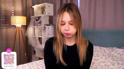 Media: Video of a young woman with long blonde hair, wearing a black top, sitting on a bed with floral bedding. Background features a white shelf, a lamp, and a pink \"Candy Crush\" logo.