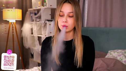 Media: Video of a blonde woman with fair skin and a long, straight mane, wearing a black top, blowing smoke into the air in a cozy bedroom with a white lamp, shelves, and floral bedspread.