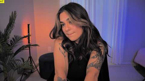 Media: Video of a Latina woman with long, wavy hair, dark skin, and a sleeve tattoo, smiling in a modern living room with orange and blue lighting, green plants, and a black armchair.